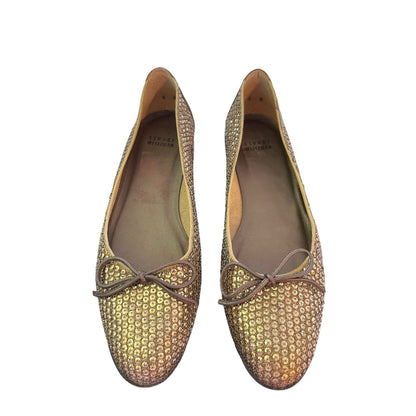 A pair of flats covered in gold-toned sequins with a leather bow on the front, shown against a plain white background.