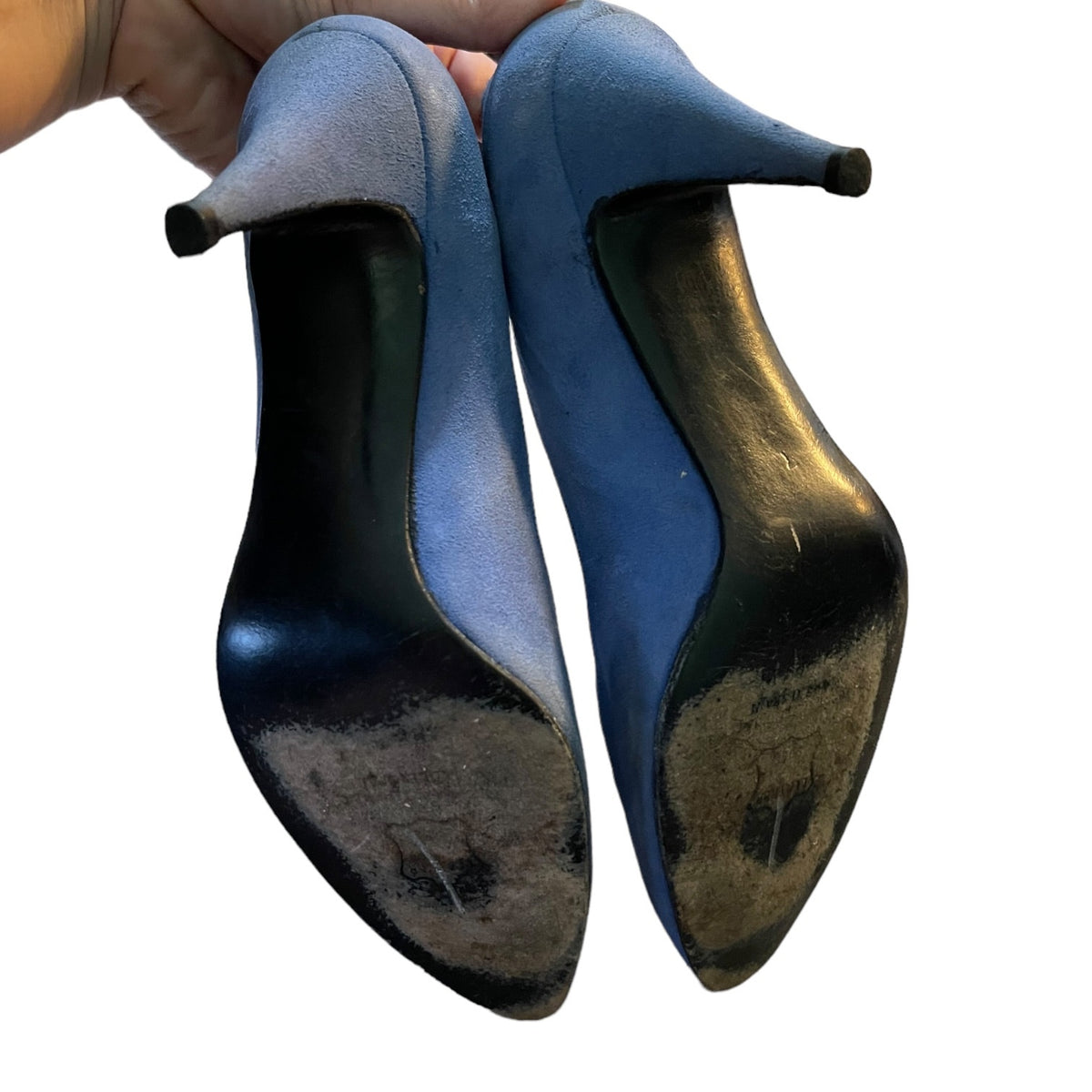 A pair of women's pointed-toe high heels in blue suede, shown from the bottom to reveal heavily scuffed black soles, held by a hand against a white background.