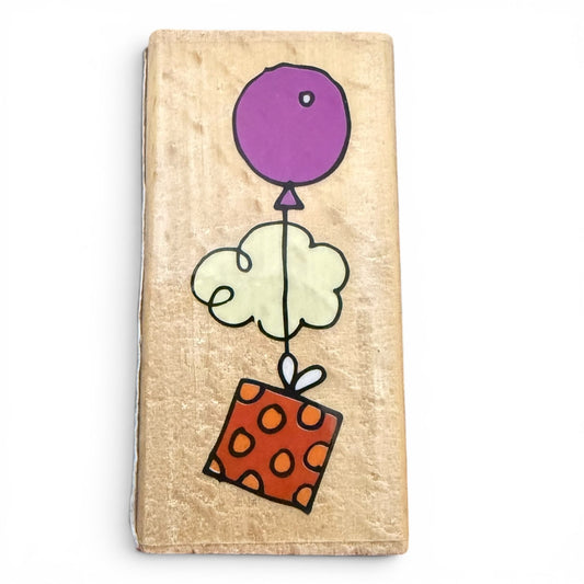Wooden block with colorful drawing of a purple balloon, white cloud, and orange box with black dots on a beige background.