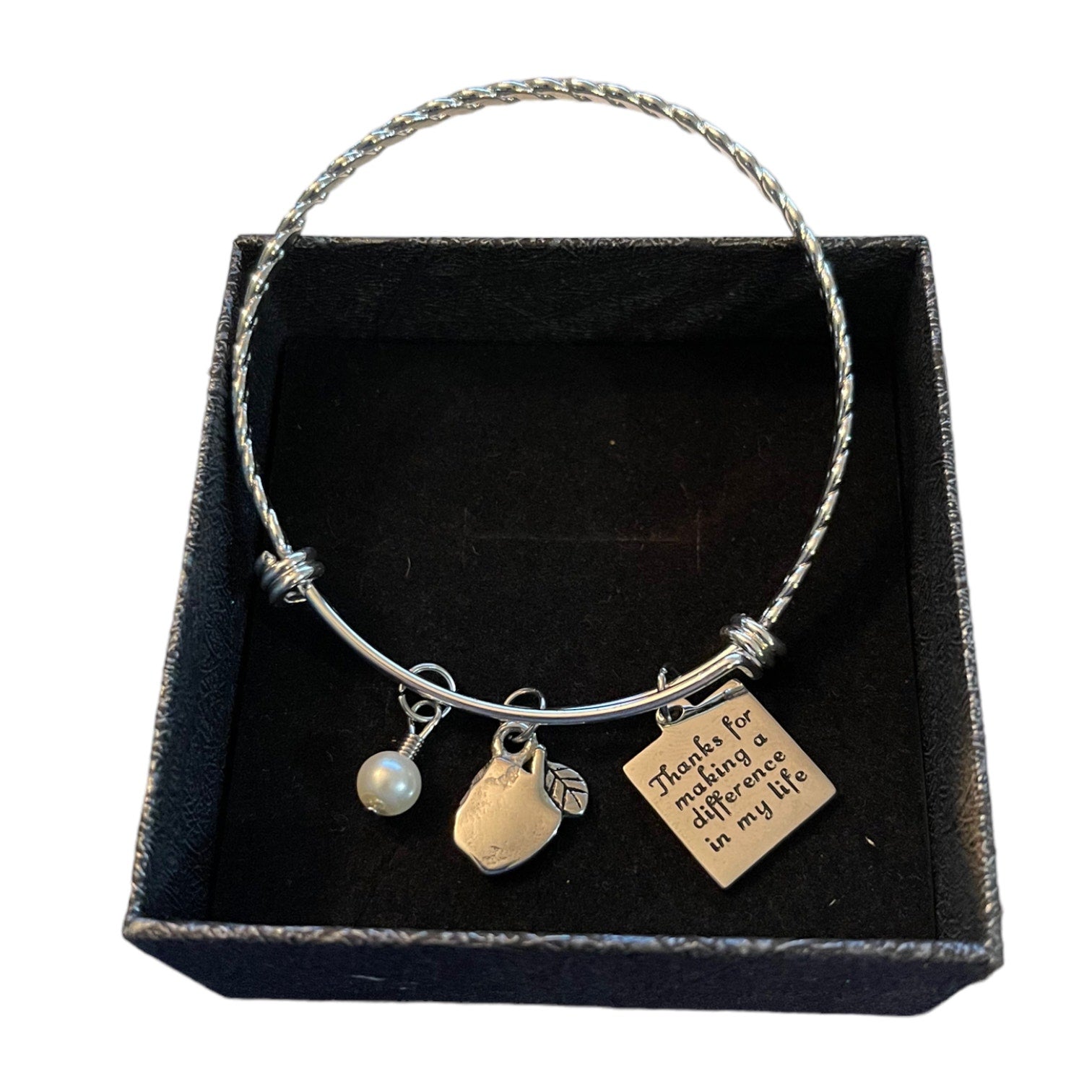 A silver-toned bangle bracelet displayed inside a black presentation box. The bracelet has three charms: a small white pearl bead, apple shaped charm, and a square engraved tag that reads, 'Thanks for making a difference in my life.