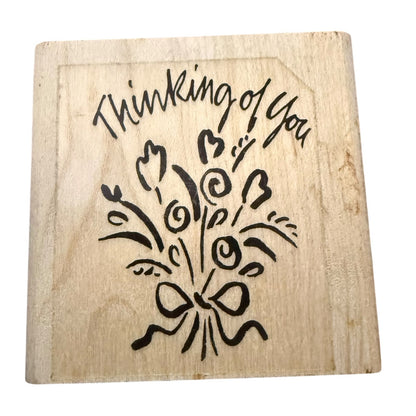 A square wooden stamp with a design that reads 'Thinking of You' above a bouquet of flowers and a bow, shown on a plain white background.