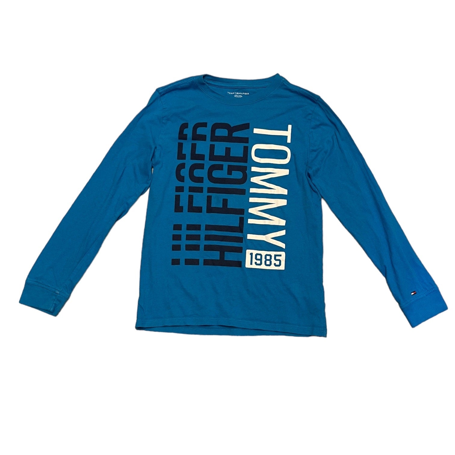 A blue long-sleeve crewneck t-shirt by Tommy Hilfiger, featuring a large vertical graphic print on the front with 'HILFIGER TOMMY 1985' in black and white.