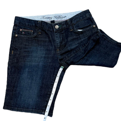 Dark wash blue denim shorts with a white and blue striped inner waistband showing the Tommy Hilfiger logo, displayed on a white background with a measuring tape draped along the inseam.