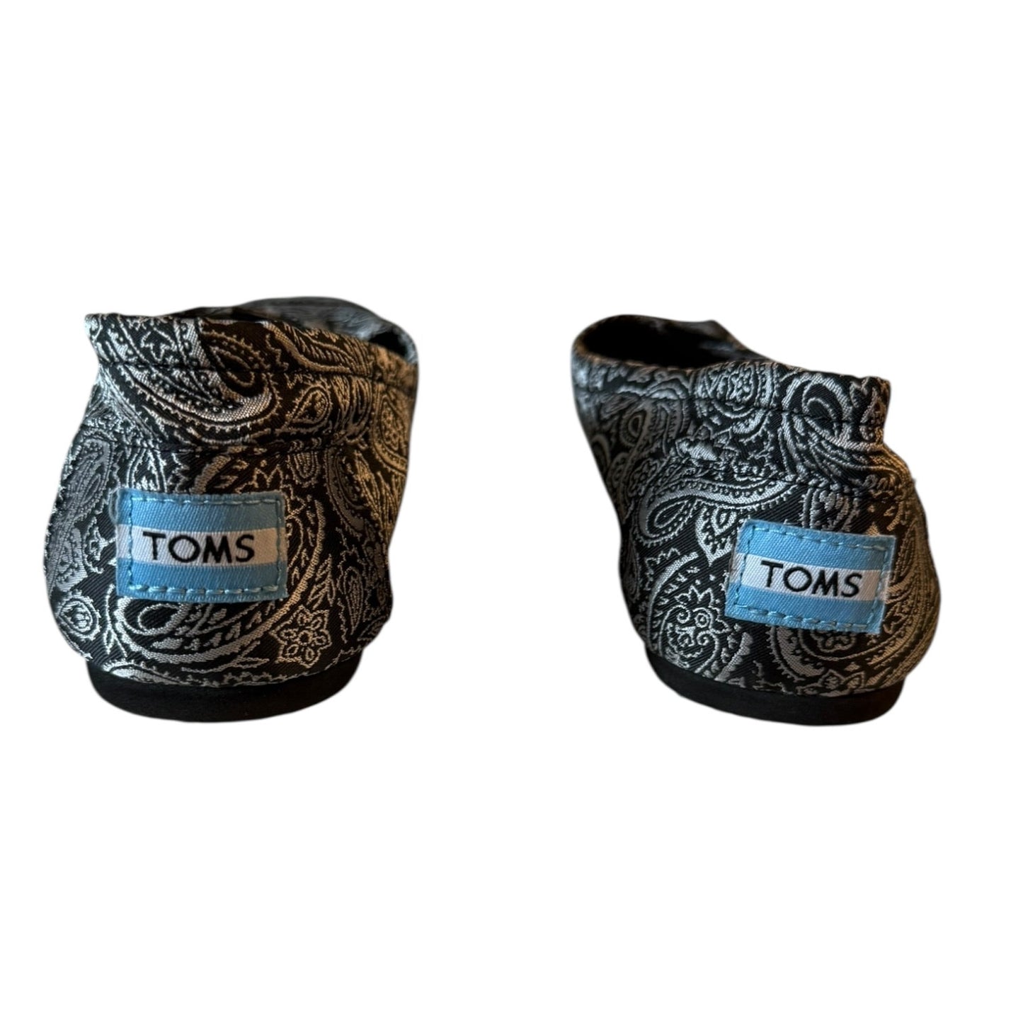 A pair of TOMS slip-on shoes with a black and silver paisley pattern and a light blue brand patch on the side, shown against a plain white background.
