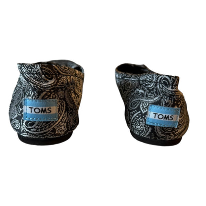 A pair of TOMS slip-on shoes with a black and silver paisley pattern and a light blue brand patch on the side, shown against a plain white background.
