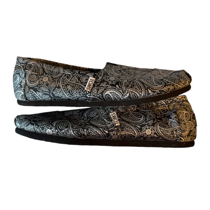 A pair of slip-on flat shoes with a black and silver paisley pattern, displayed against a plain white background.
