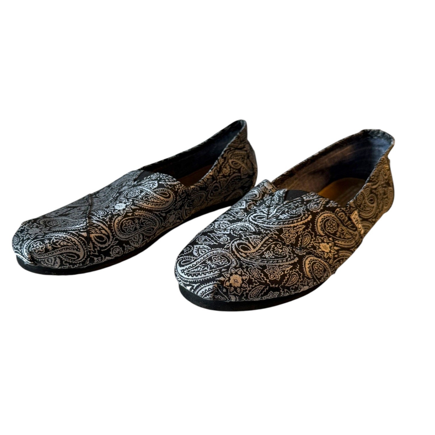 A pair of slip-on loafers with an intricate black and silver paisley pattern, displayed against a plain white background.