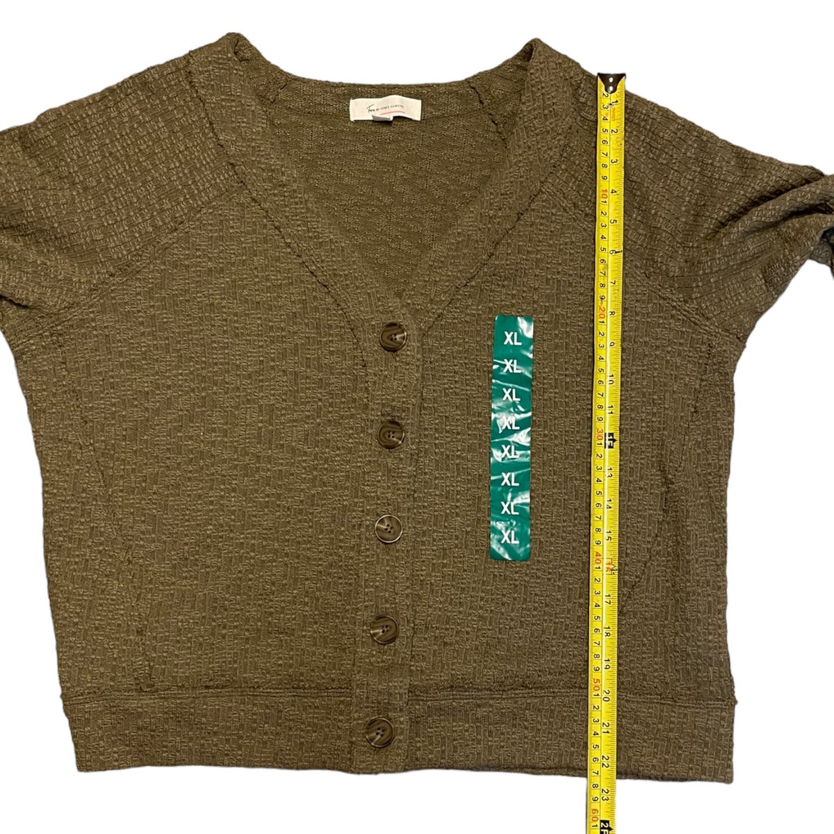An green knit cardigan sweater laid flat on a white background, shown with a yellow tape measure along the right side for size reference. The cardigan has a V-neck, short sleeves, and front buttons.