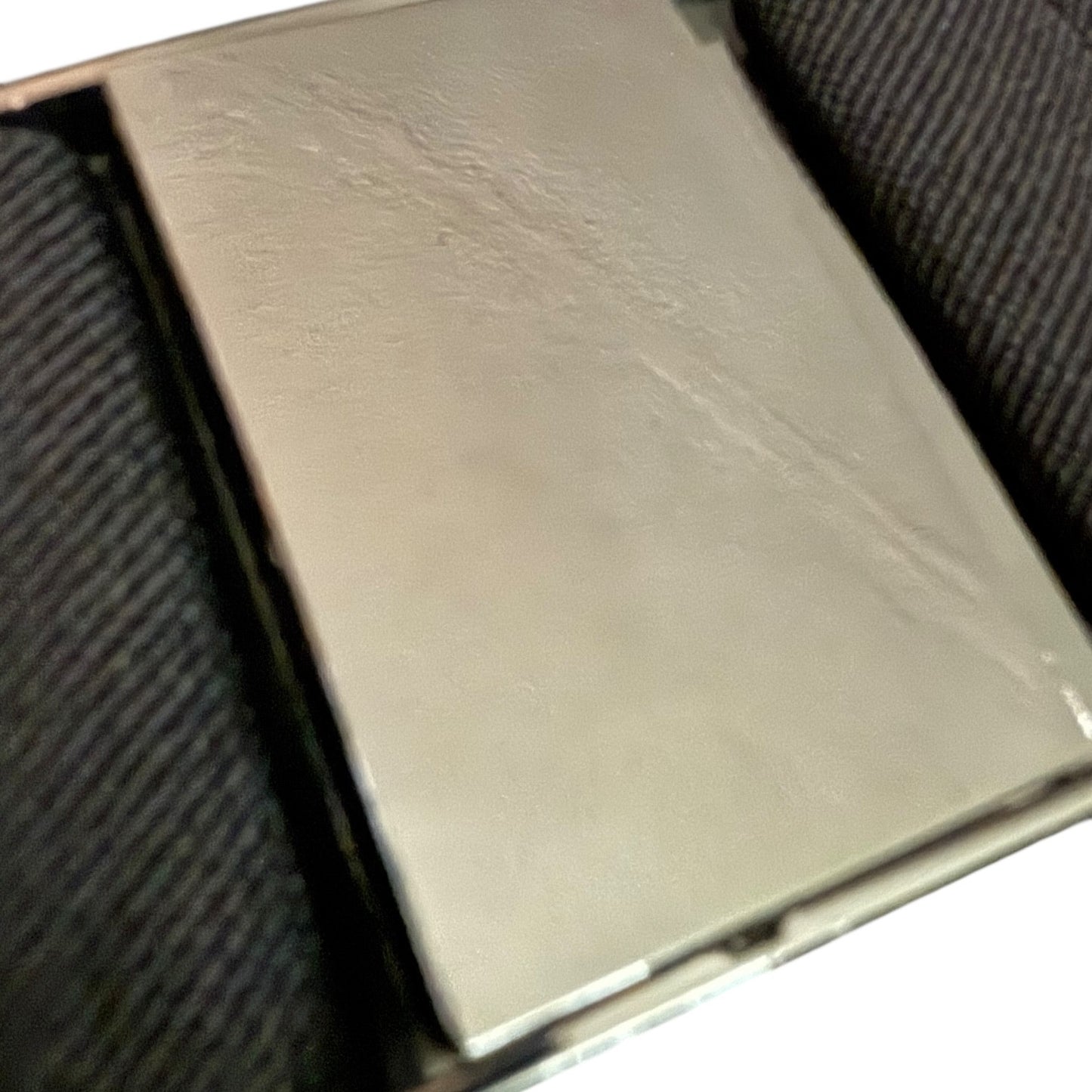 A close-up of a metallic silver rectangular belt buckle, resting on a dark textured fabric surface.
