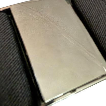 A close-up of a metallic silver rectangular belt buckle, resting on a dark textured fabric surface.