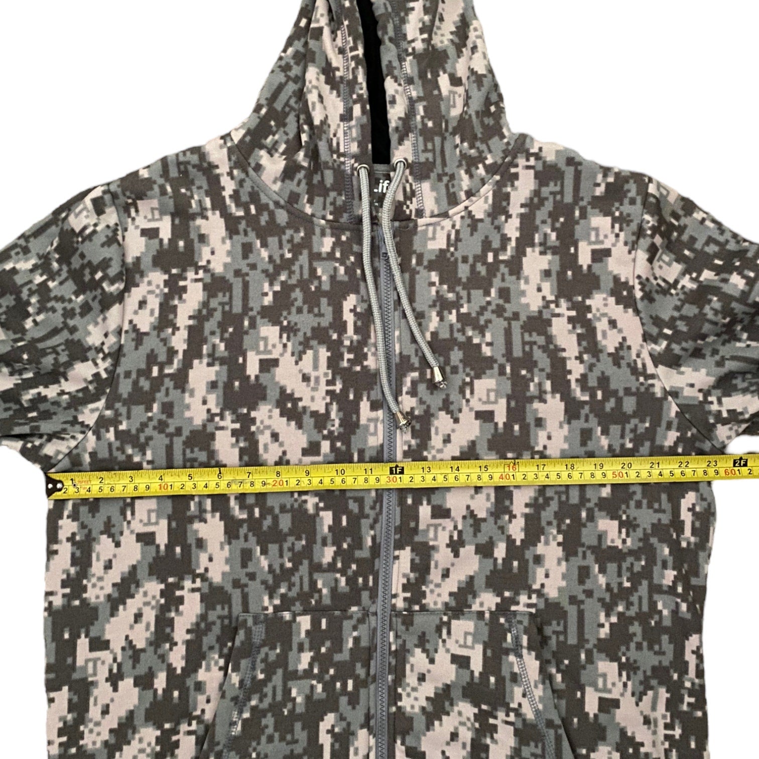 A hooded zip-up jacket with a digital camouflage pattern, shown with a yellow tape measure stretched horizontally across the chest to indicate size.