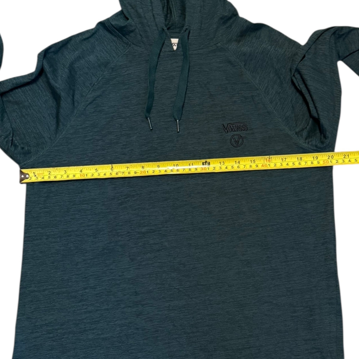 A dark green hooded shirt with a yellow tape measure stretched horizontally across the chest to show the width. The hoodie features a small  Vans logo on the left chest.