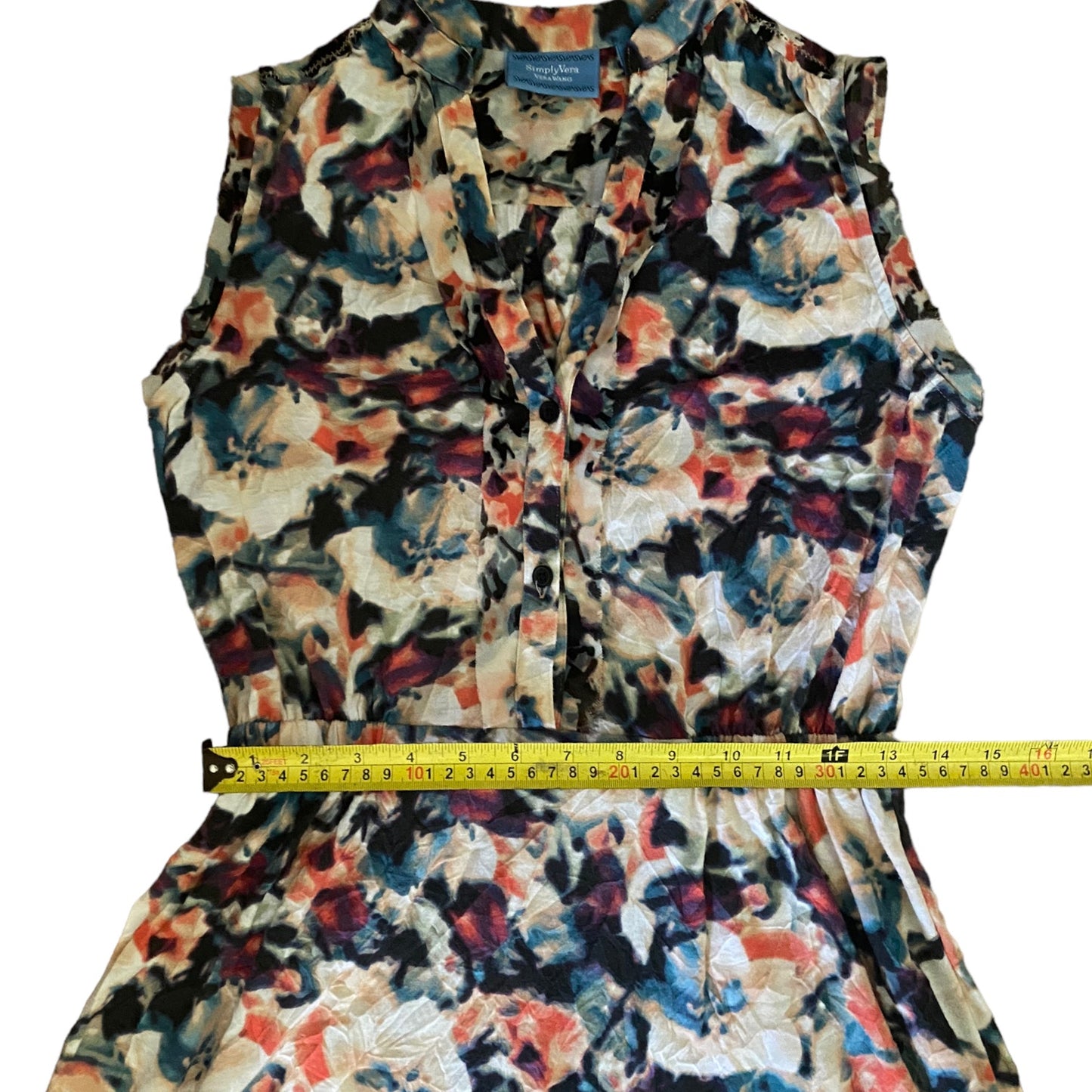 A sleeveless floral print dress with a V-neckline and ruffled shoulders, shown with a yellow tape measure wrapped around the waist to indicate measurements. The dress features an abstract watercolor-style pattern in black, blue, red, and beige tones.