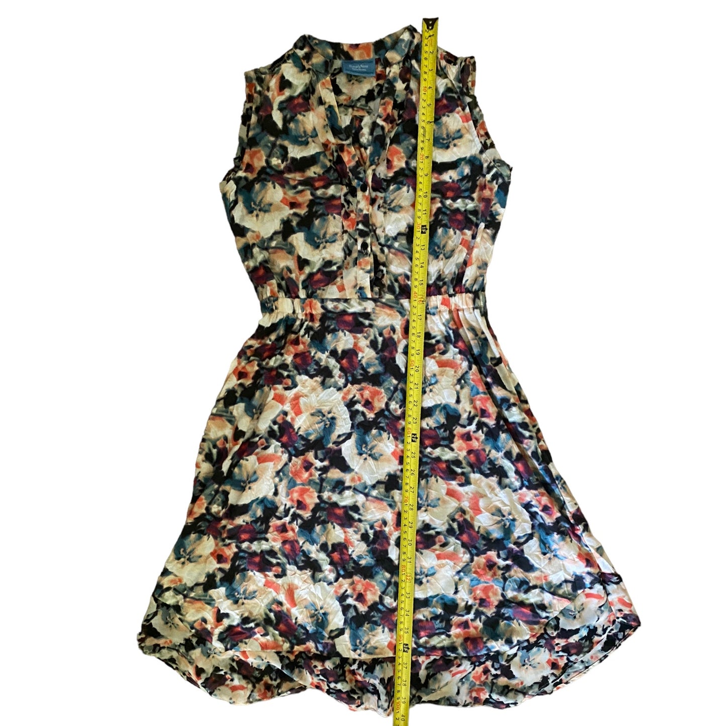 A sleeveless floral print dress with a v neckline , shown flat against a white background with a yellow measuring tape running vertically down the center.