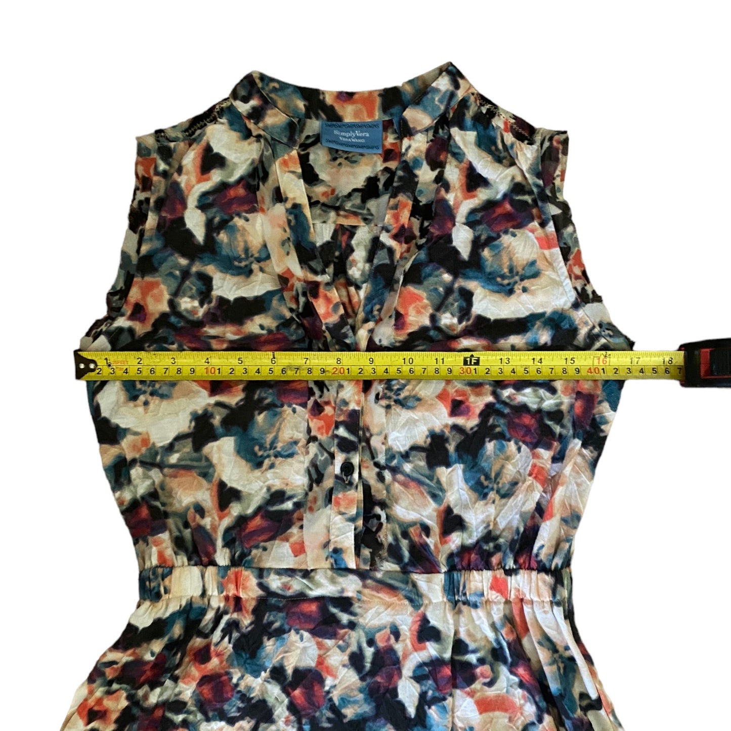 A sleeveless floral dress with a multicolor abstract print in blue, red, black, and beige, shown with a yellow tape measure stretched across the chest to indicate width.