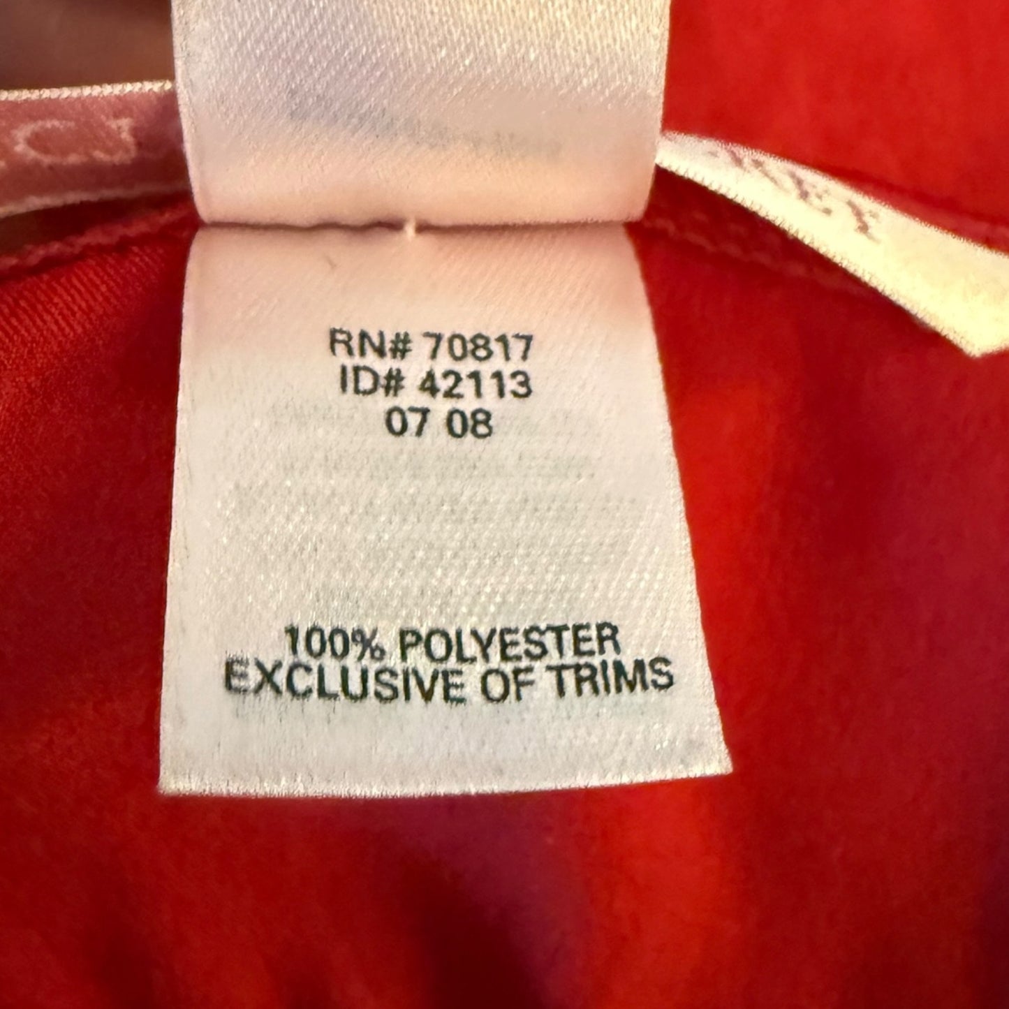 Close-up of a white fabric care label sewn onto a red garment, showing printed text including '100% POLYESTER EXCLUSIVE OF TRIMS'.