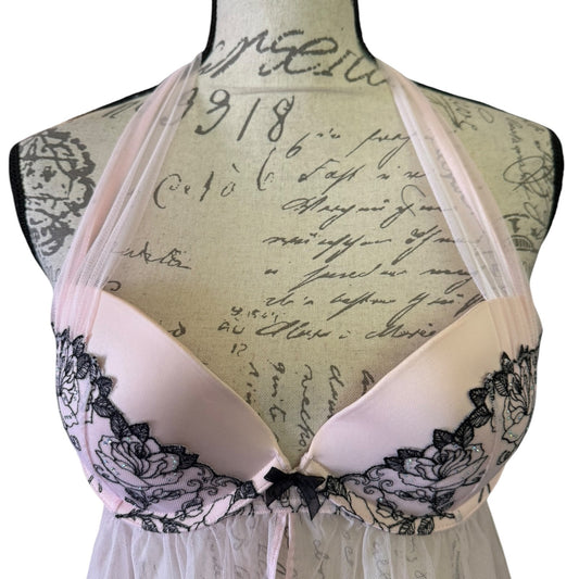 A pale pink halter-style lingerie with black floral lace trim and a small black bow at the center, displayed on a vintage-style mannequin torso with cursive script print.