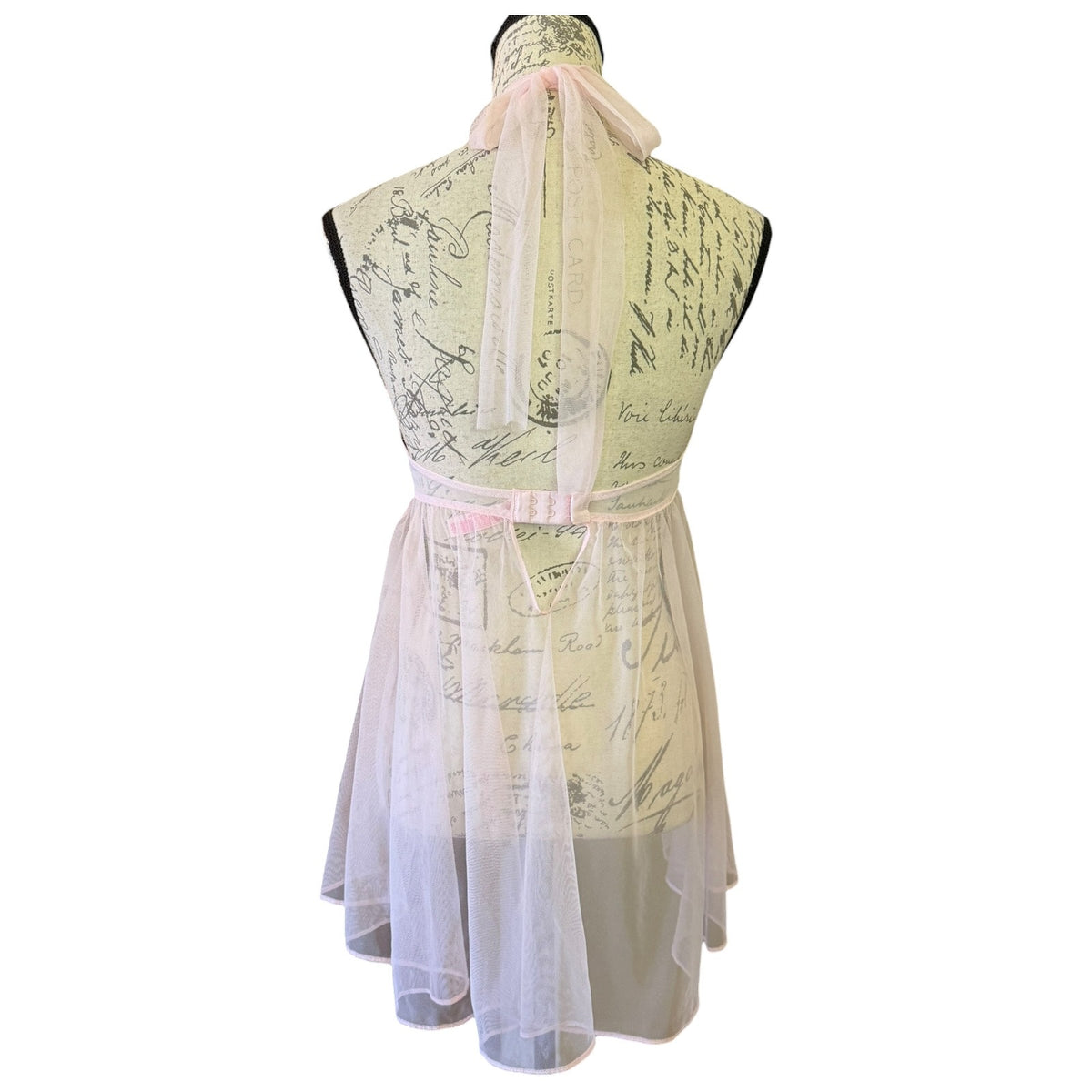 A sleeveless babydoll-style lingerie dress displayed on a mannequin, featuring a cream-colored printed bodice with cursive script, a light pink ribbon belt at the waist, and a sheer white tulle skirt.