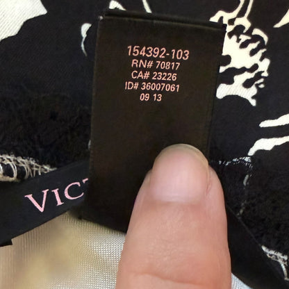 A close-up of a black garment tag held by a finger, showing printed product codes and a partial Victoria's Secret logo on the fabric.