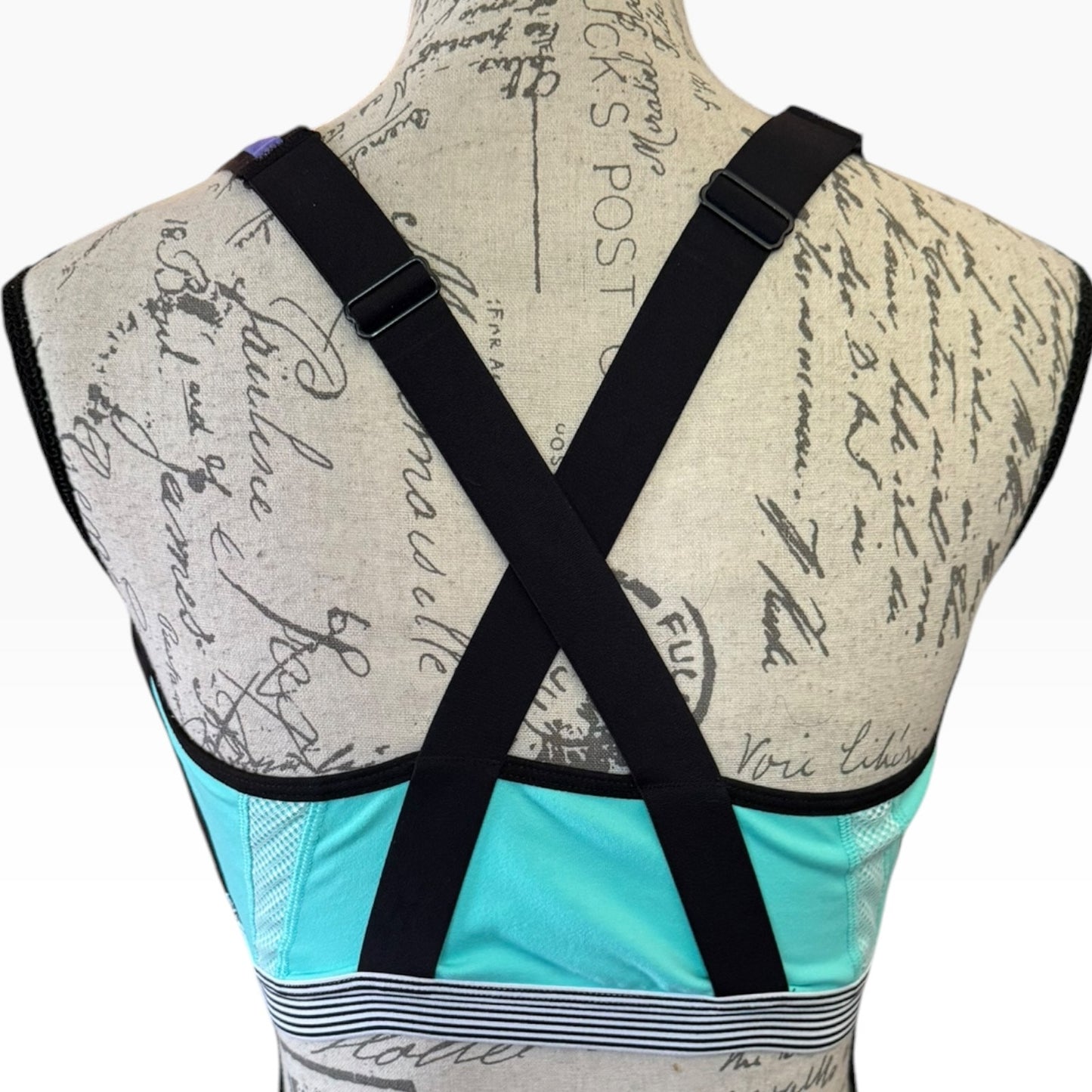 Back view of a sports bra on a patterned dress form, featuring black adjustable shoulder straps that cross in an X shape and a blue band at the bottom.