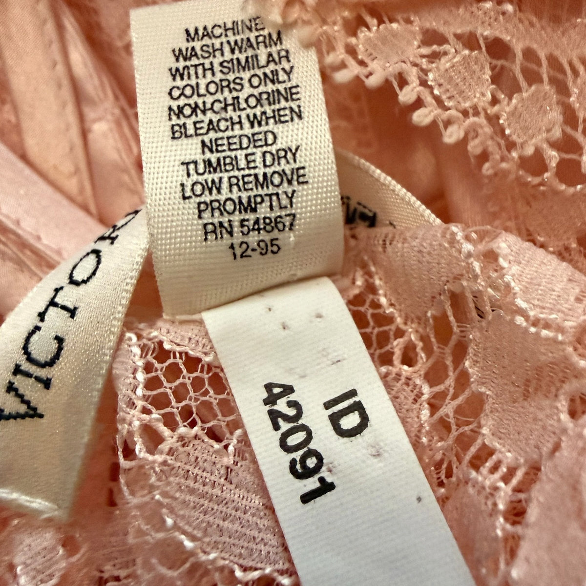 Close-up of a garment's interior labels showing a Victoria brand tag and a size/ID label, set against pink lace fabric.