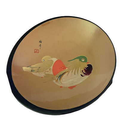 Round plate with a glossy gold-toned glaze, featuring a hand-painted traditional Japanese-style illustration of two ducks. The plate has a black rim and a small black calligraphy seal on the left side.