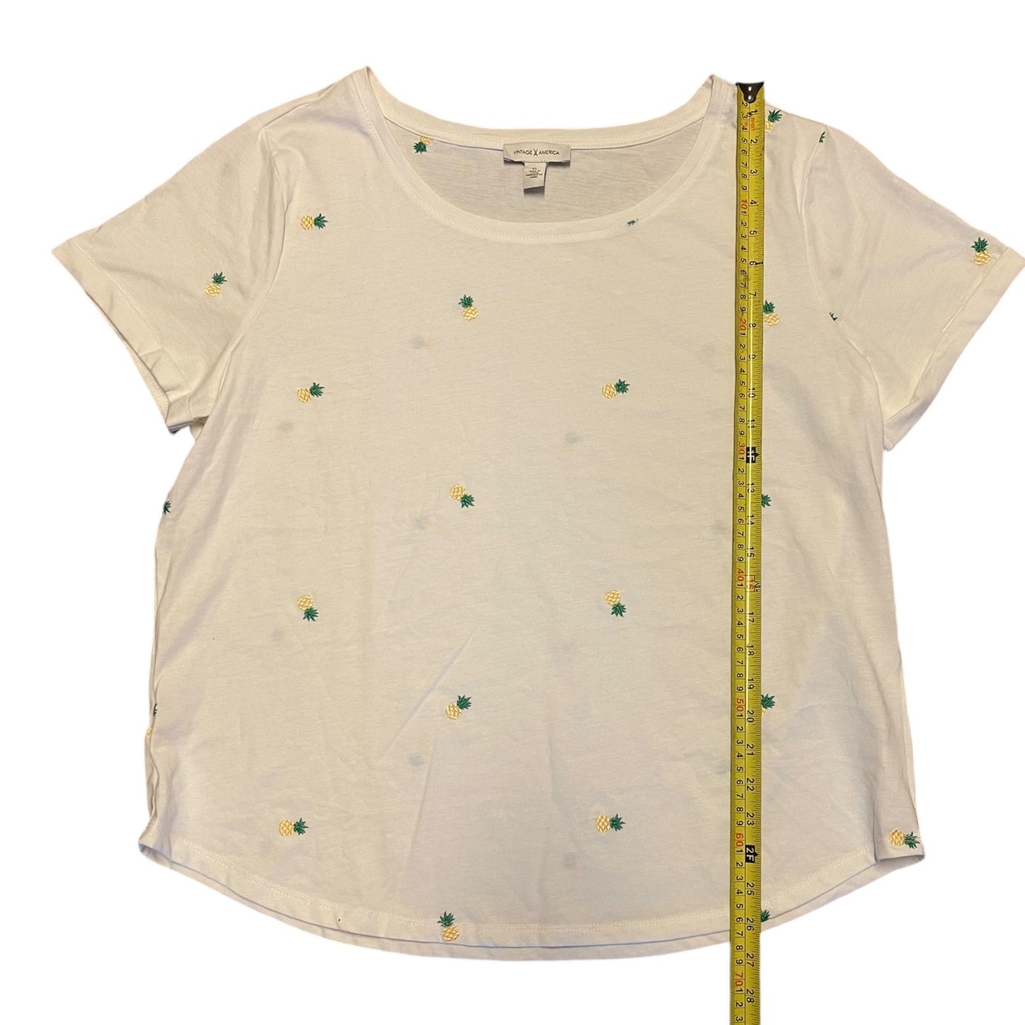 An white short-sleeve t-shirt with small green and yellow pineapples, shown flat with a yellow measuring tape standing vertically on the right side for scale.