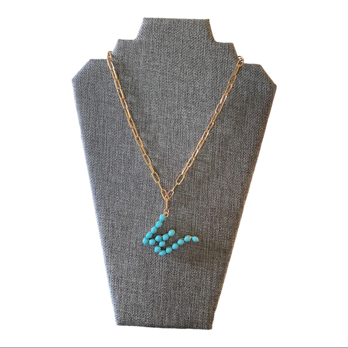 A gold chain necklace with a turquoise bead pendant, displayed on a gray fabric jewelry bust against a white background.