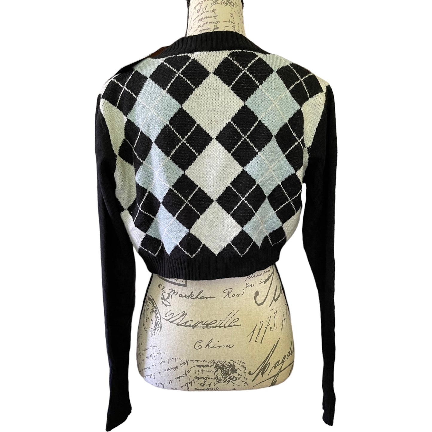 A cropped knit cardigan sweater shown from the back on a mannequin torso. The cardigan features a black and cream argyle pattern with light blue-gray diamonds and solid black sleeves and ribbed collar.
