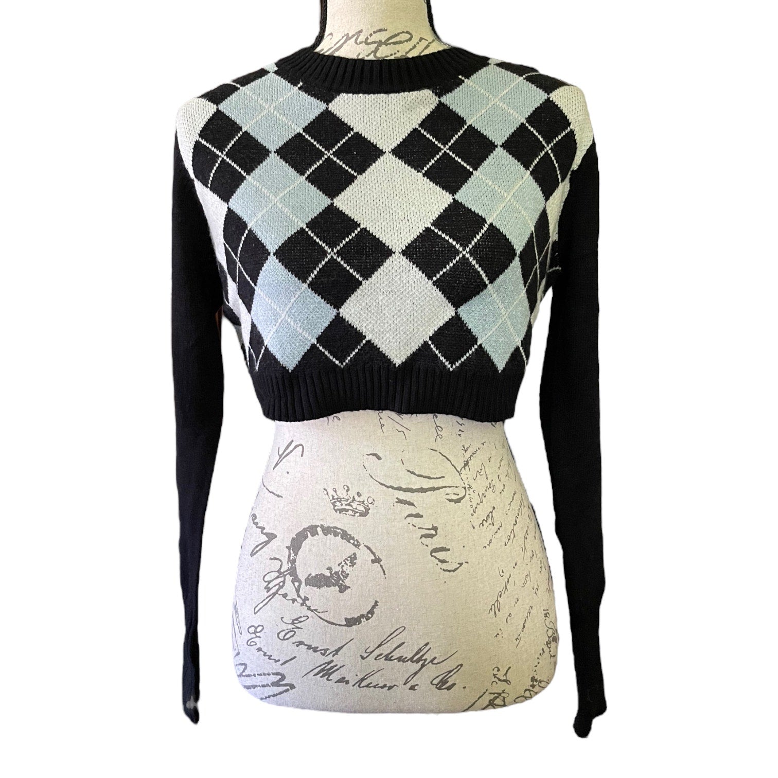 A cropped long-sleeve knit sweater displayed on a mannequin torso. The sweater features a black, off-white, and light blue argyle pattern across the chest and shoulders, with solid black sleeves and a black ribbed crew neck and hem.