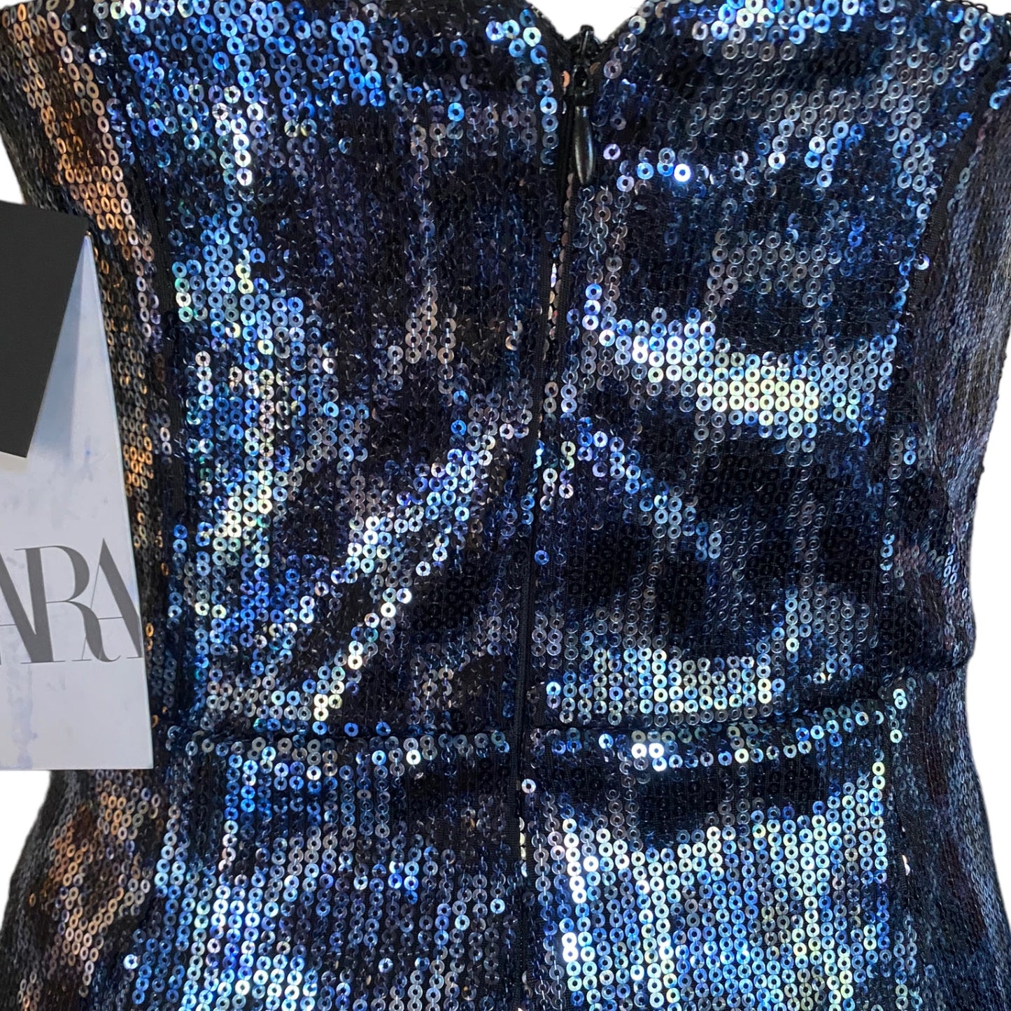 Close-up of a sleeveless sequined dress  with a back zipper, covered in iridescent blue, navy, and silver sequins. A partial paper tag is visible on the left side.