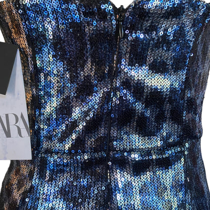 Close-up of a sleeveless sequined dress  with a back zipper, covered in iridescent blue, navy, and silver sequins. A partial paper tag is visible on the left side.